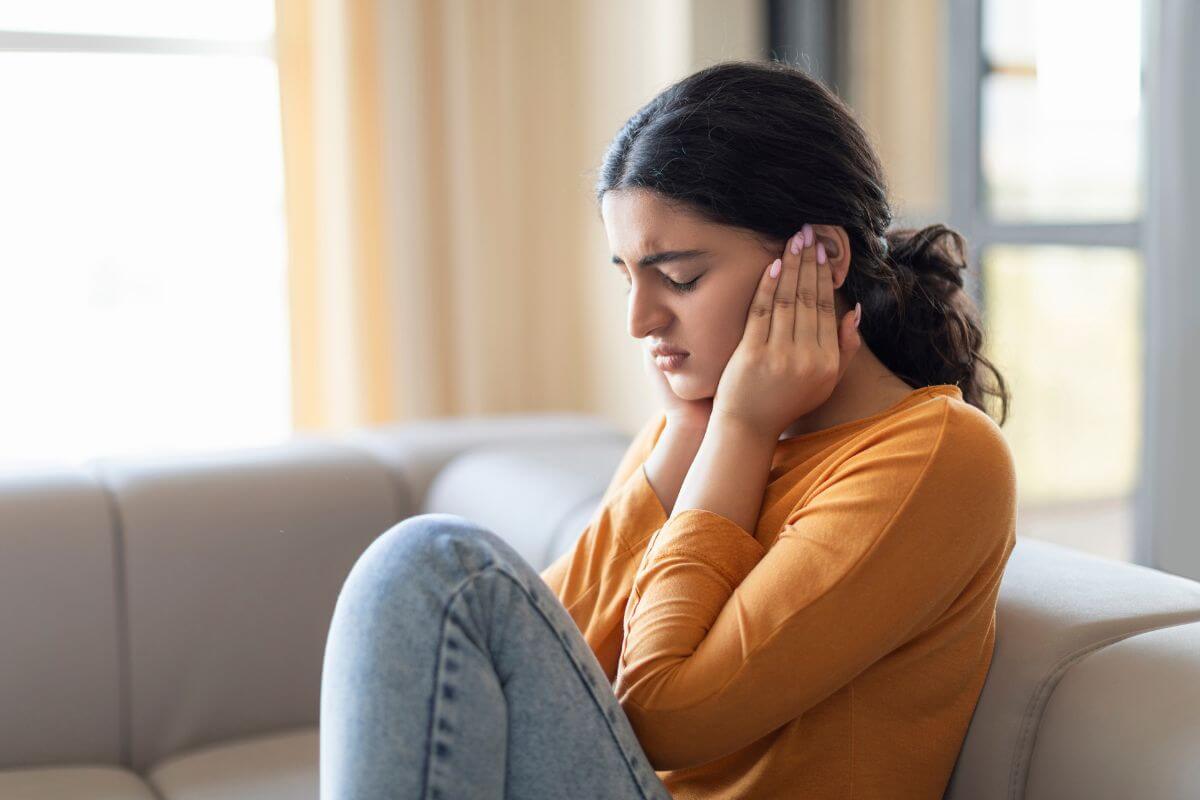 Ratgeber Frau sitzt angespannt auf dem Sofa und hält sich die Ohren – Sinnbild für plötzliche Angst und innere Alarmreaktionen ohne erkennbaren Auslöser