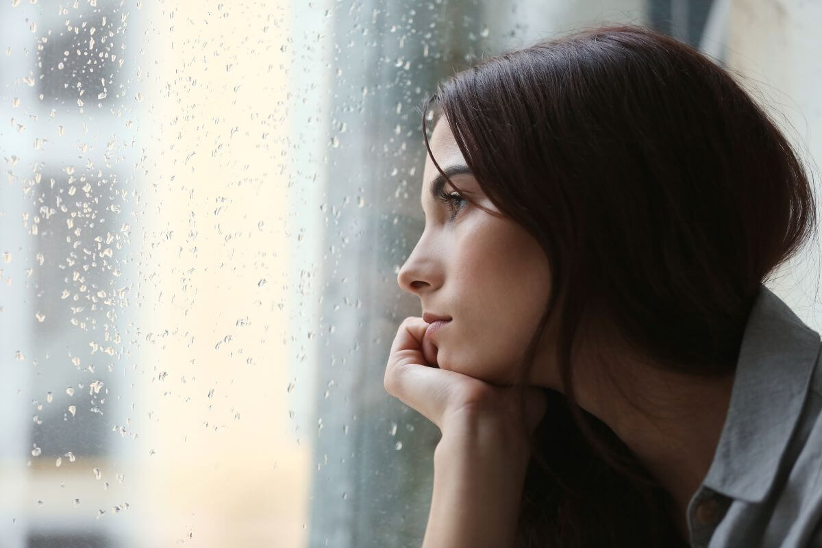Ratgeber Junge Frau schaut nachdenklich aus dem Fenster an einem Regentag – ein Symbol für sozialen Rückzug, Überforderung und das Bedürfnis nach innerer Ruhe.